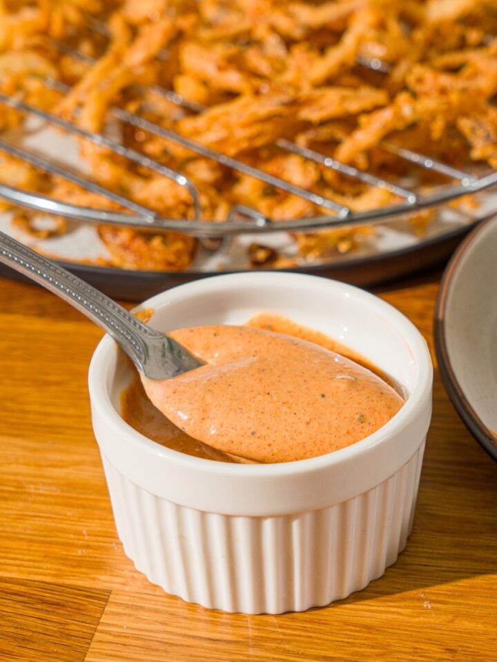 A white bowl with Whataburger creamy pepper sauce. There is a spoon in the bowl. The bowl is on a wooden counter with a plate of crispy onions.