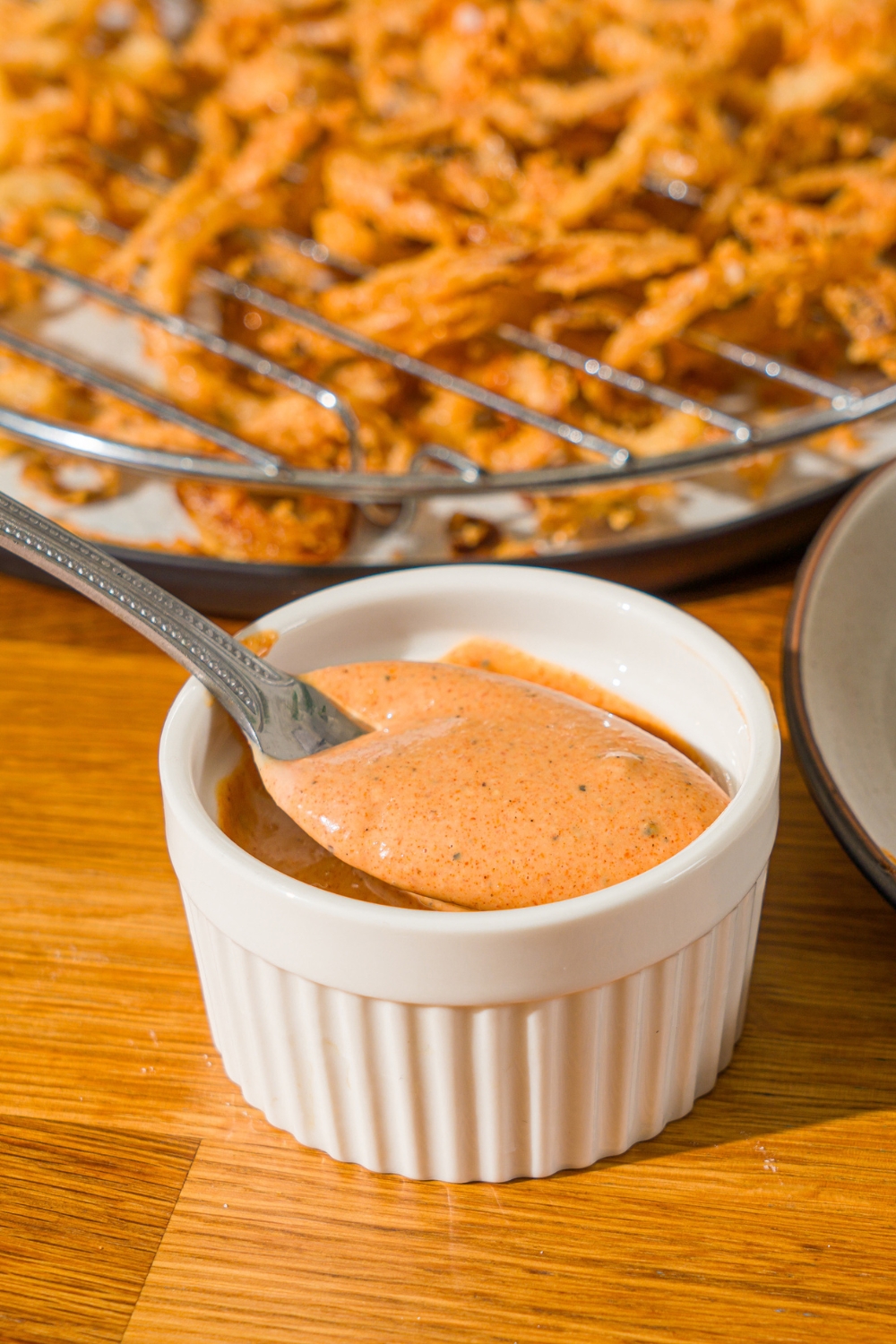 A white bowl with Whataburger creamy pepper sauce. There is a spoon in the bowl. The bowl is on a wooden counter with a plate of crispy onions.