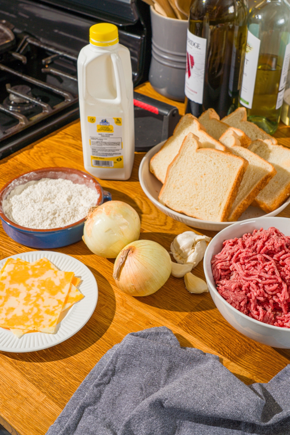 A wooden counter with several bowls in various sizes containing ingredients to make Whataburger patty melt including ground beef, flour, sliced Monterey Jack cheese, onion, buttermilk, and sliced bread.