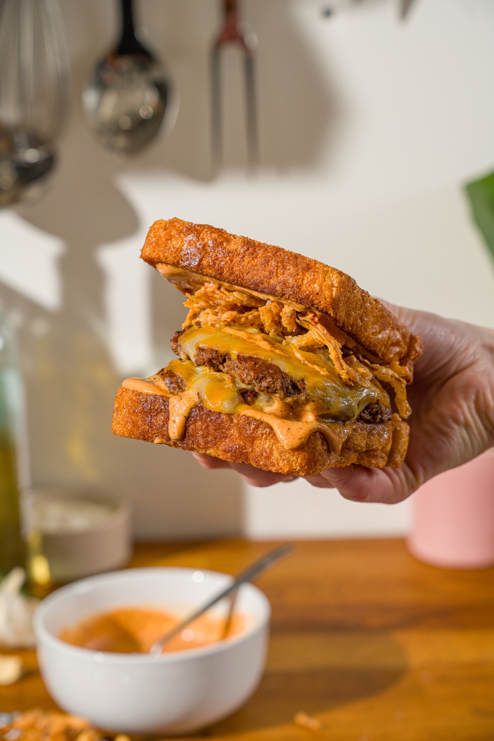 A hand holding a copycat Whataburger patty melt. There is a bowl of Whataburger patty melt sauce on a wooden counter blurred in the background.