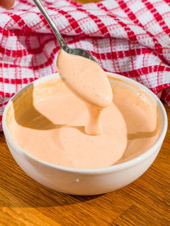 A spoon with comeback sauce on it, drizzling some of the comeback sauce into a white bowl filled with the sauce.