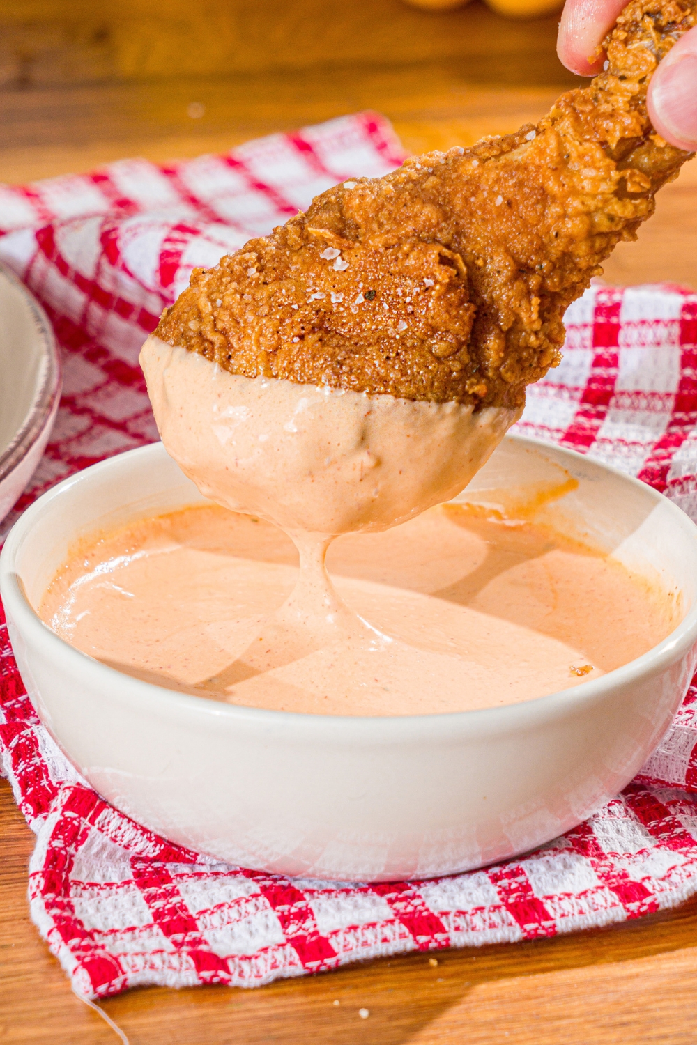 A fried chicken drumstick with comeback sauce on it being held over a white bowl that is filled with the comeback sauce.