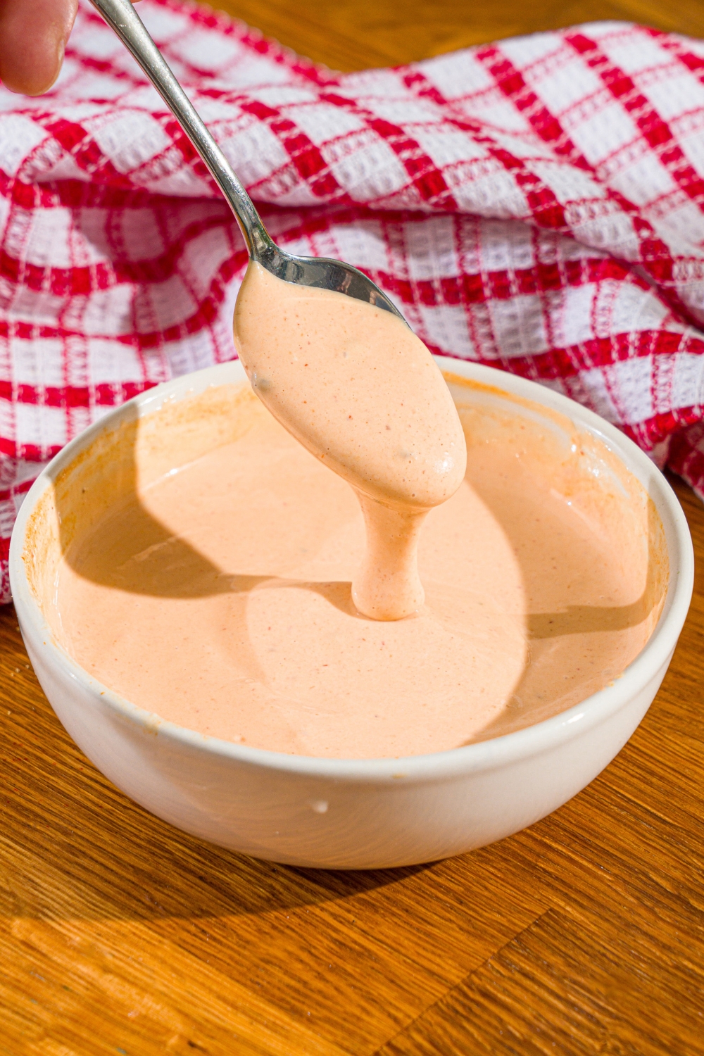 A spoon with comeback sauce on it, drizzling some of the comeback sauce into a white bowl filled with the sauce.