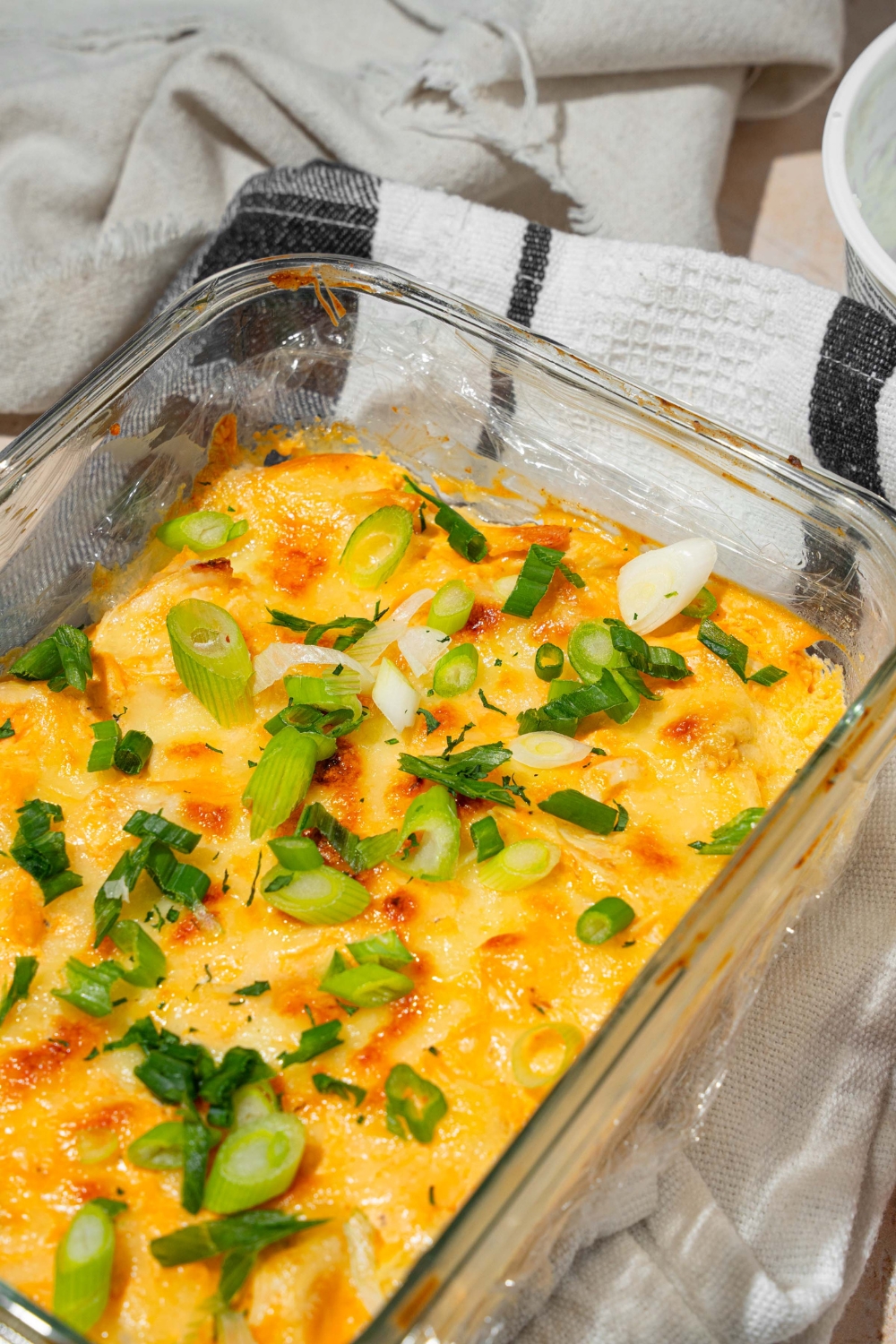 A glass baking dish with cottage cheese Buffalo chicken dip garnished with sliced green onion. The dish is on a dish towel on a tan counter with a small plate of garnishes.