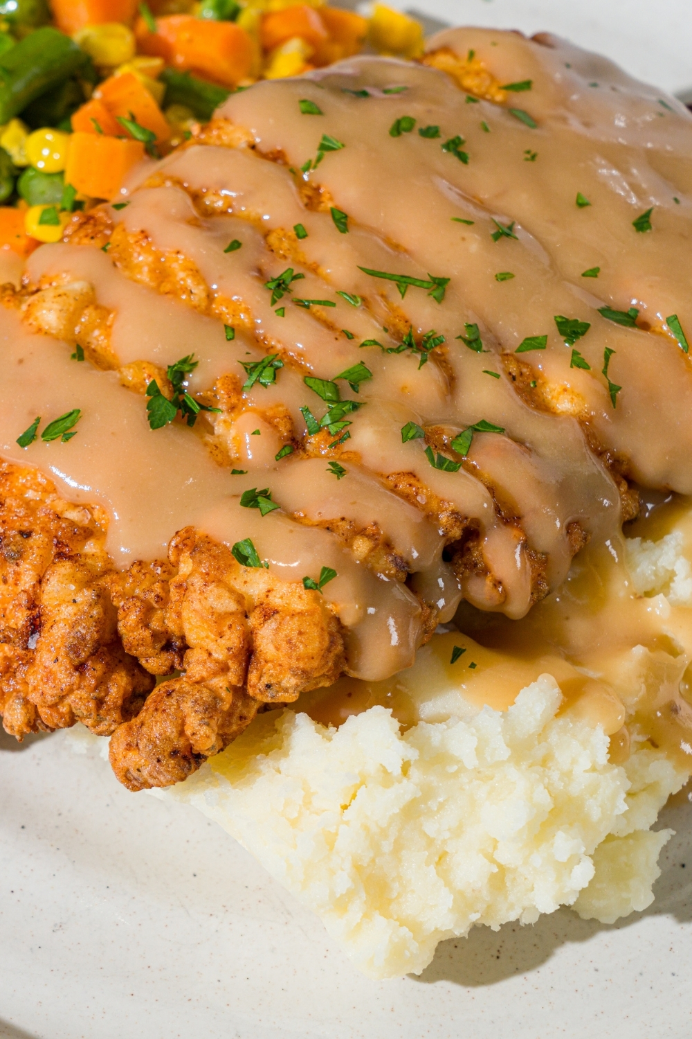 A white plate with country fried chicken drizzled with gravy and served over mashed potatoes and mixed vegetables. The chicken is garnished with fresh parsley.
