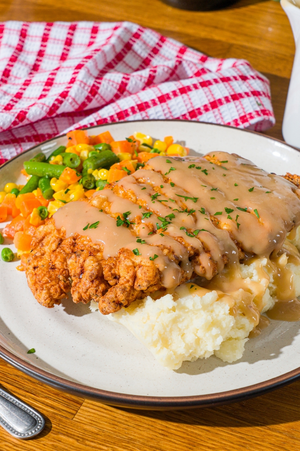A white plate with country fried chicken drizzled with gravy and served over mashed potatoes and mixed vegetables. The chicken is garnished with fresh parsley. The plate is on a wooden counter with a red striped napkin.