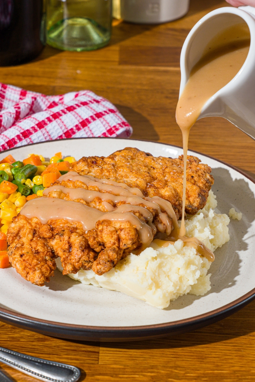 A white plate with country fried chicken served over mashed potatoes and mixed vegetables. Gravy is being drizzled over the chicken. The plate is on a wooden counter with a red striped napkin.