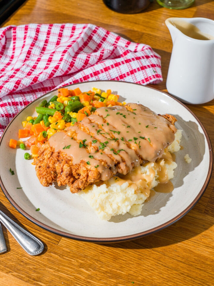 A white plate with country fried chicken drizzled with gravy and served over mashed potatoes and mixed vegetables. The chicken is garnished with fresh parsley. The plate is on a wooden counter with a red striped napkin.