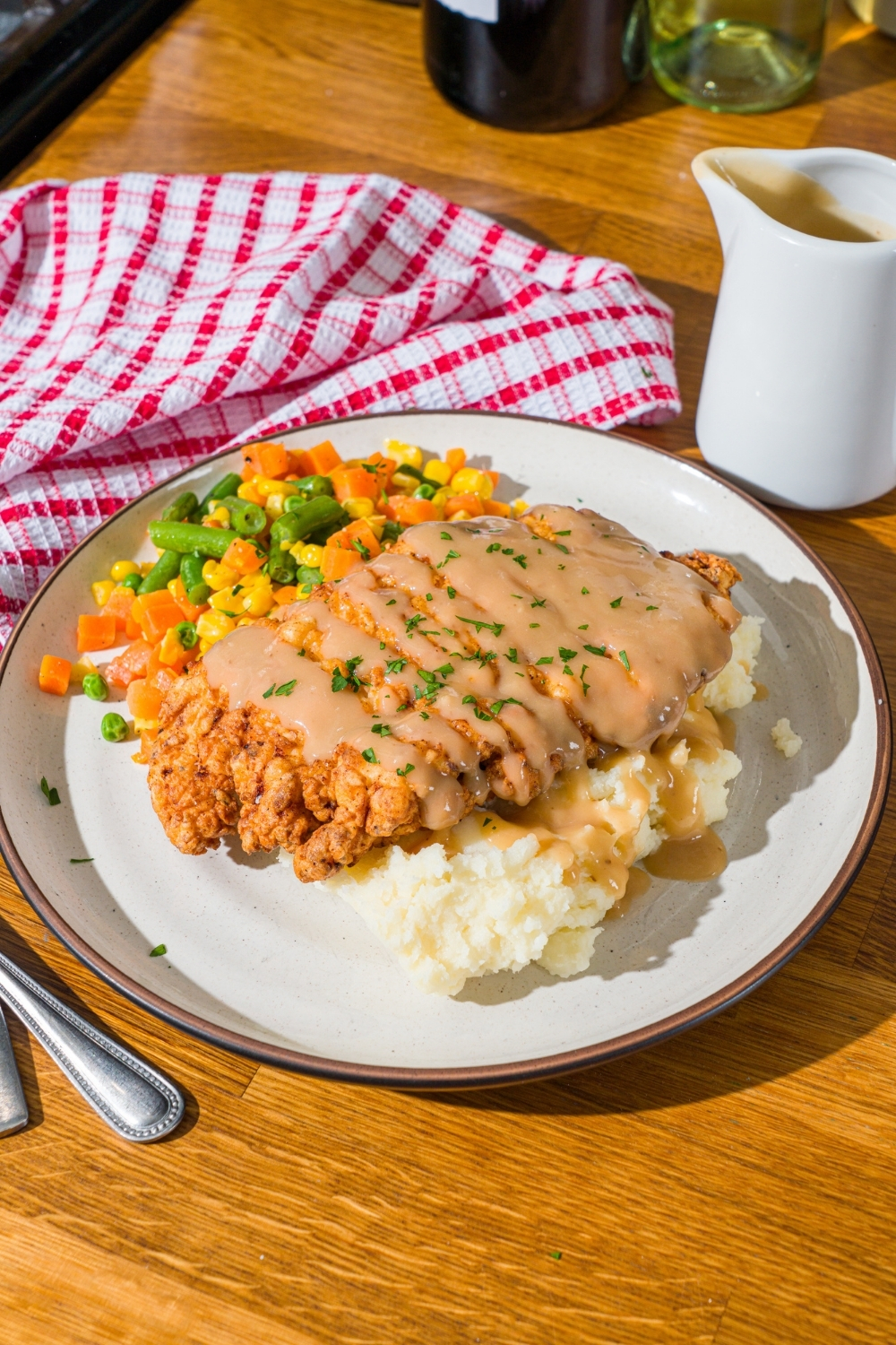 A white plate with country fried chicken drizzled with gravy and served over mashed potatoes and mixed vegetables. The chicken is garnished with fresh parsley. The plate is on a wooden counter with a red striped napkin.