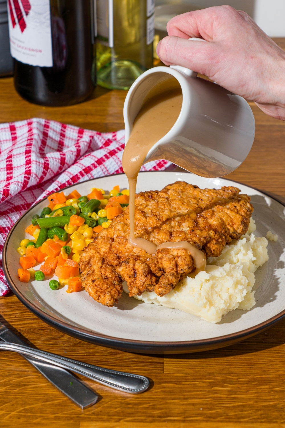 A white plate with country fried chicken served over mashed potatoes and mixed vegetables. Gravy is being drizzled over the chicken. The plate is on a wooden counter with a red striped napkin.