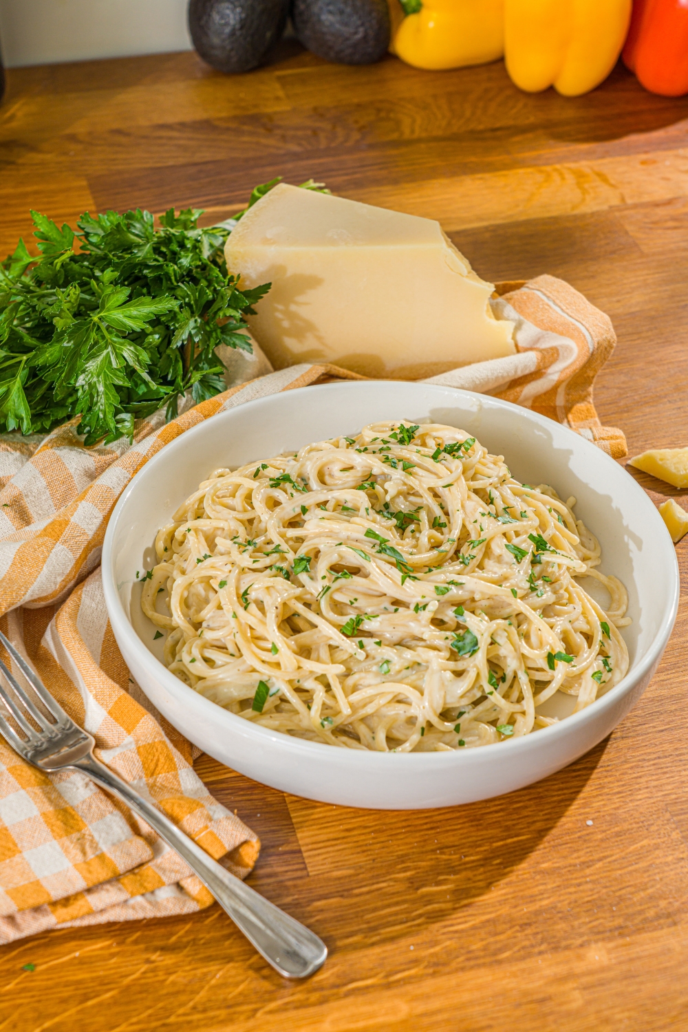 A white bowl with spaghetti tossed in a creamy garlic parmesan sauce garnished with fresh parsley. A fork is taking a bite of pasta from the bowl. The bowl is on a wooden counter with a fork and yellow checkered napkin.