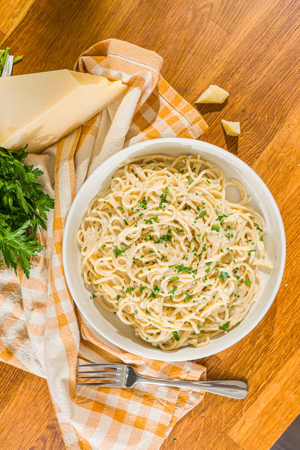 A white bowl with spaghetti tossed in a creamy garlic parmesan sauce garnished with fresh parsley. A fork is taking a bite of pasta from the bowl. The bowl is on a wooden counter with a fork and yellow checkered napkin.