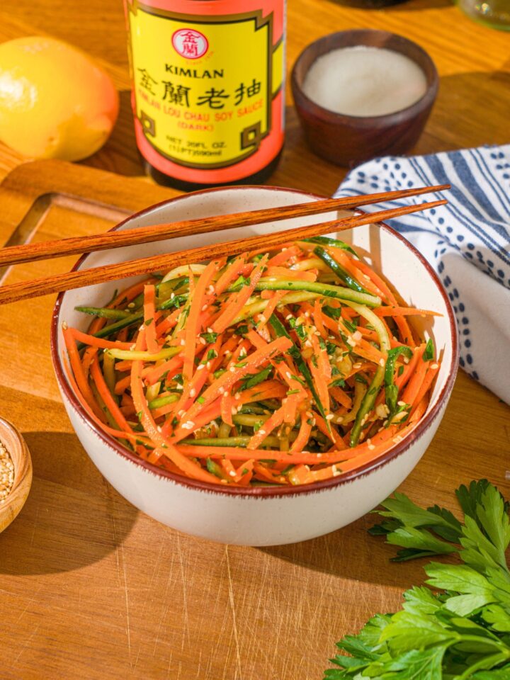 A bowl of thinly sliced cucumber and carrot salad in a white bowl with chop sticks resting on top.