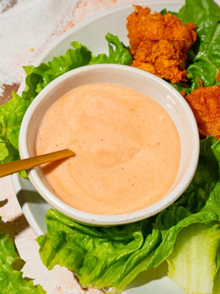 A spoon submerged in a white bowl that is filled with Dave's hot chicken sauce. There is some lettuce and chicken tenders around the sauce on a plate.