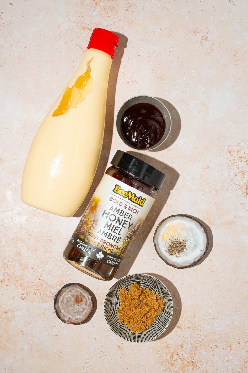 A bottle of chipotle sauce, a bottle of honey, a small bowl of barbecue sauce, a small bowl of brown sugar, a small plate of salt and pepper all on a white counter.