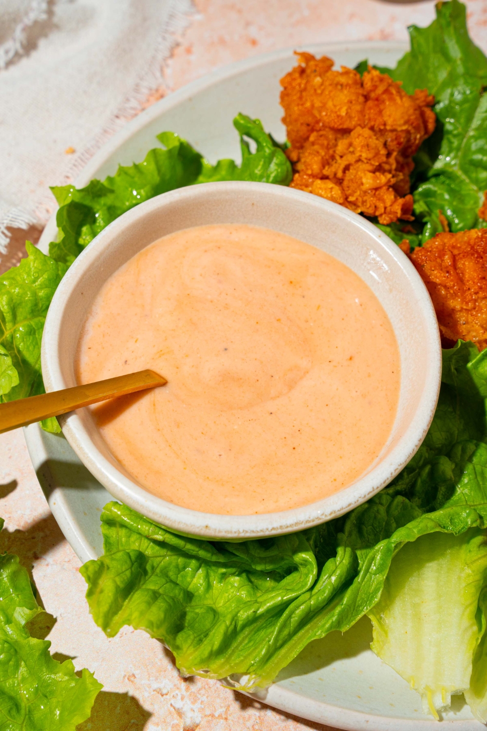 A spoon submerged in a white bowl that is filled with Dave's hot chicken sauce. There is some lettuce and chicken tenders around the sauce on a plate.