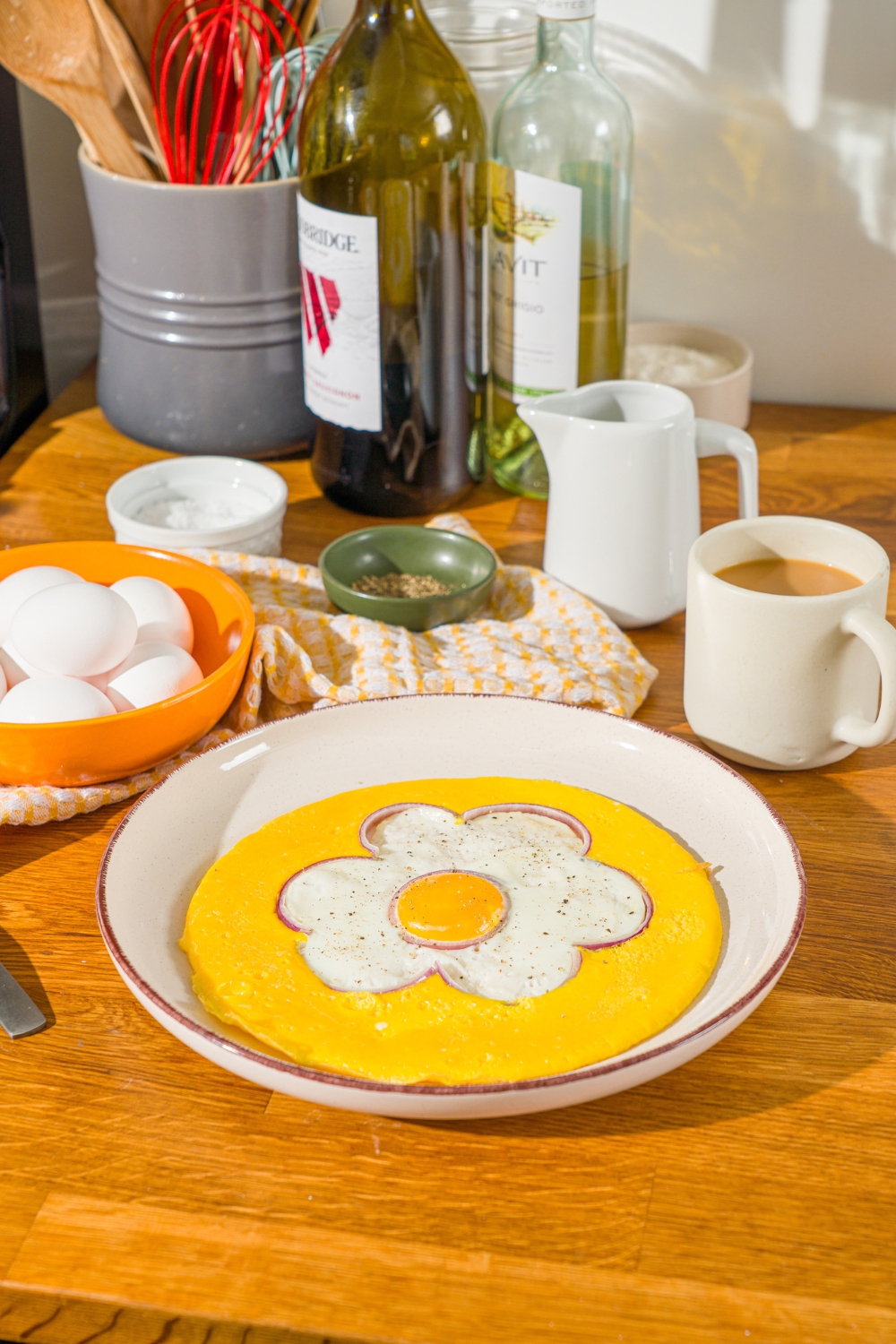 An egg flower omelette on a white plate with a bowl of eggs, a coffee cup, and a bottle of oil behind it.
