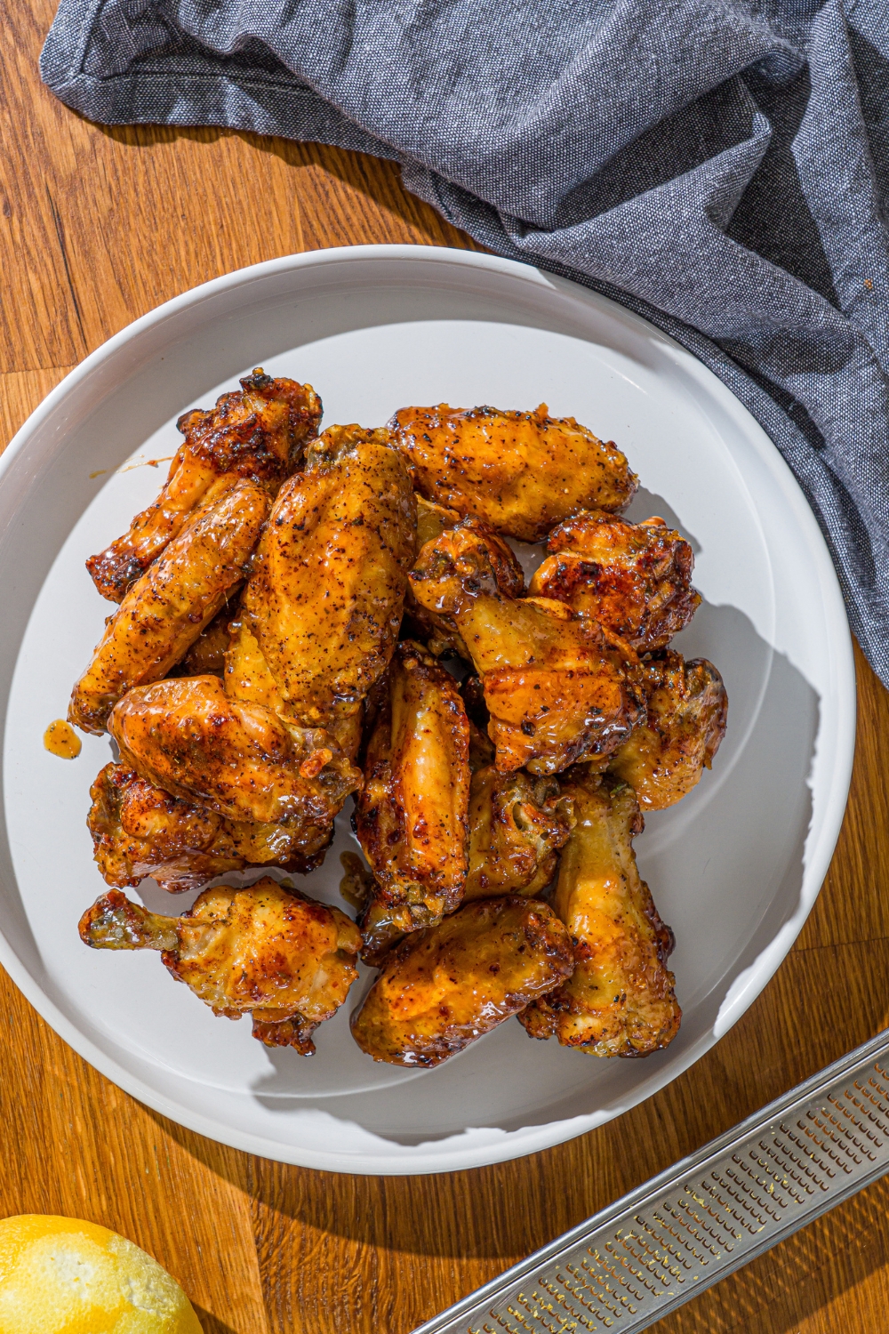 A white plate with a pile of crispy honey lemon pepper wings. The plate is on a wooden counter with a blue cloth napkin.