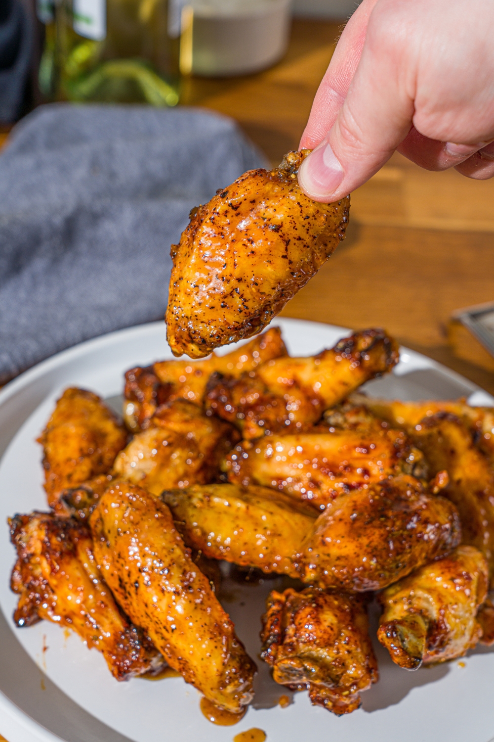 A white plate with a pile of crispy honey lemon pepper wings. A hand is holding one of the wings. The plate is on a wooden counter with a blue cloth napkin.