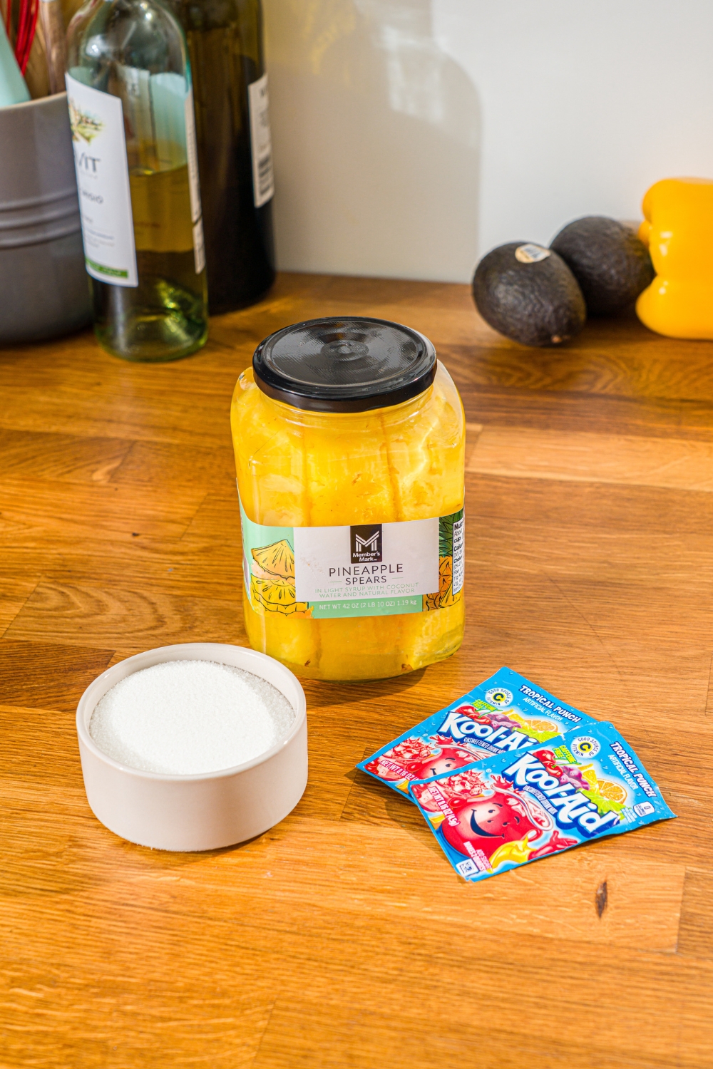 A jar of pineapple spears, a cup of sugar, and two Kool-Aid packets on a wood counter.