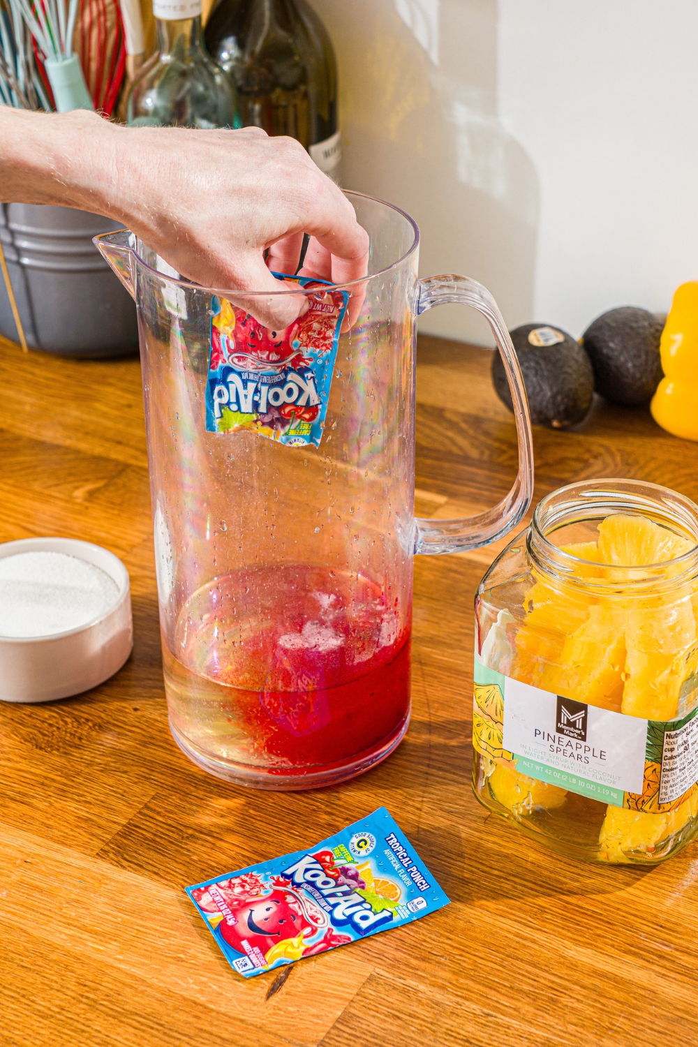 A hand pouring a red Kool-Aid packet into a glass pitcher.