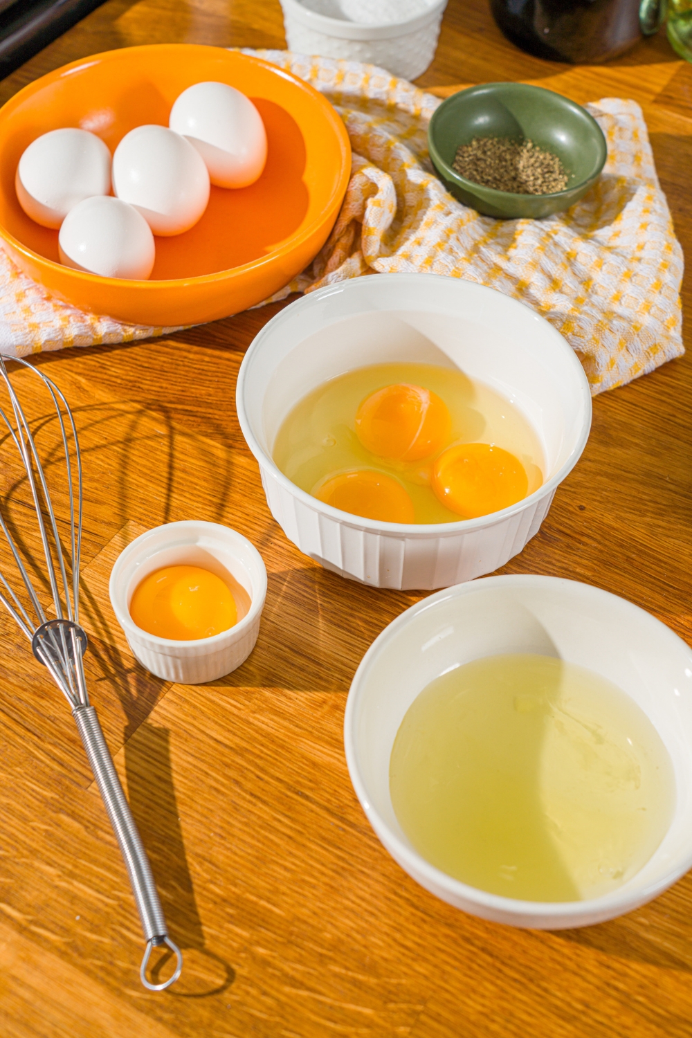 Four eggs in an orange bowl, in front of that is three eggs cracked in a white bowl, a bowl with one egg yolk, and a bowl with egg whites.