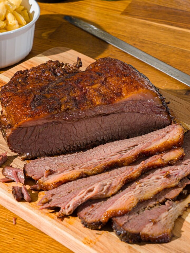 A wooden cutting board with oven baked beef brisket. Some of the brisket has been sliced. There is a bowl of mac and cheese on the board.