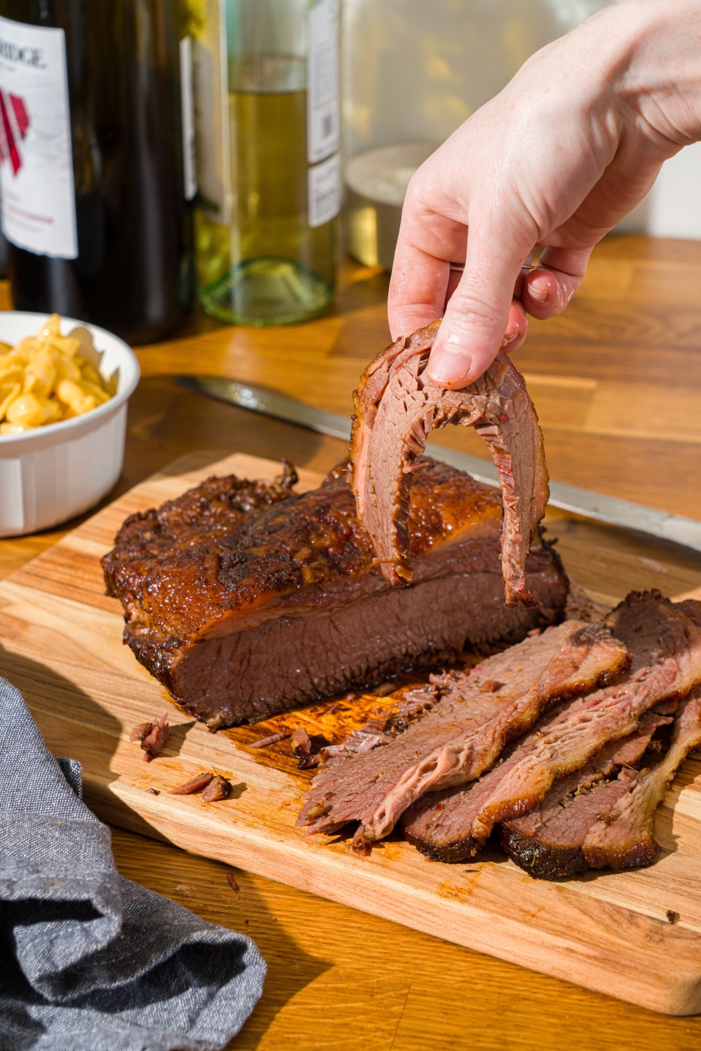 A wooden cutting board with oven baked beef brisket with several slices of brisket on the board. A hand is holding one of the slices of brisket. There is a bowl of mac and cheese on the board.