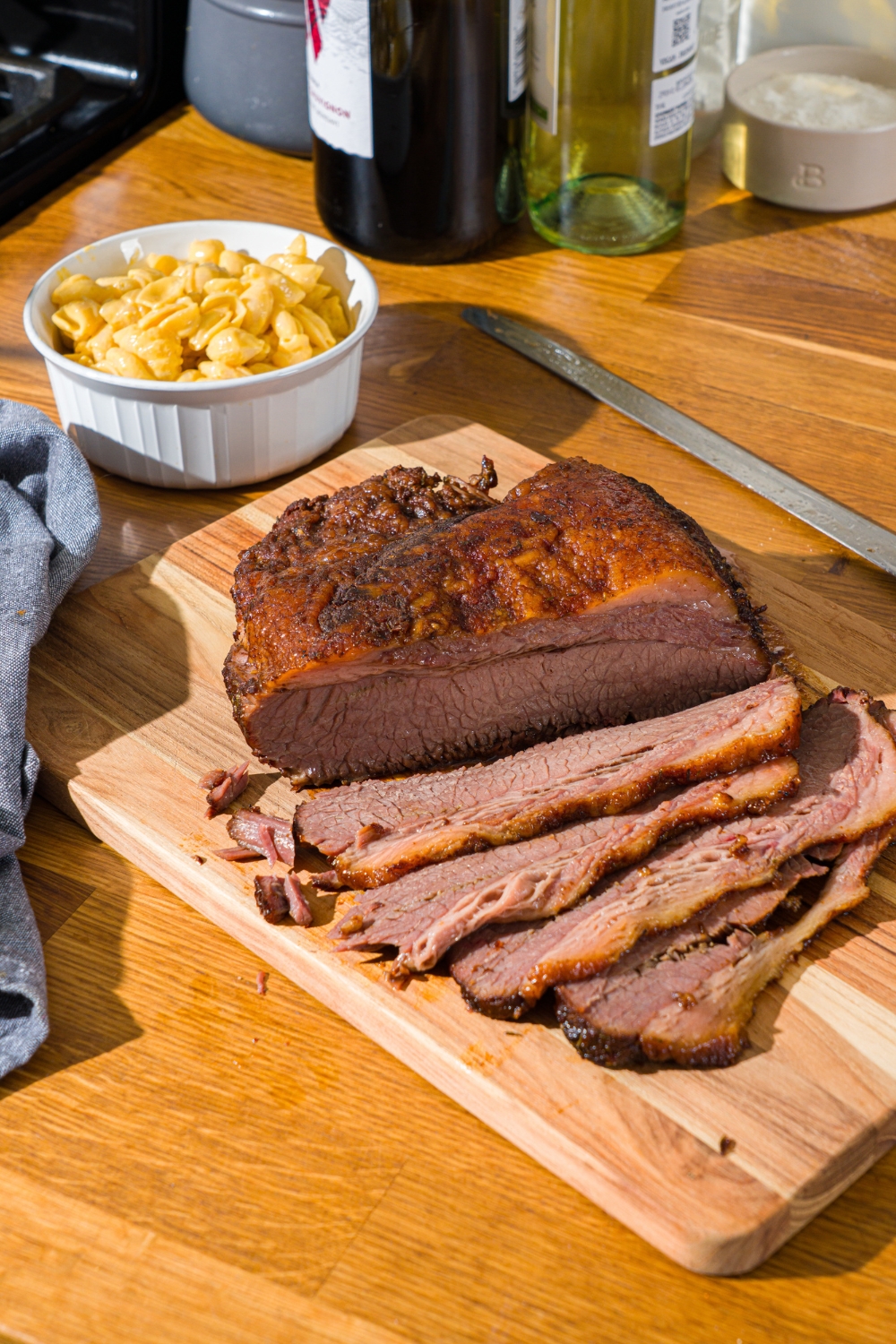 A wooden cutting board with oven baked beef brisket with several slices of brisket on the board. There is a bowl of mac and cheese on the board.