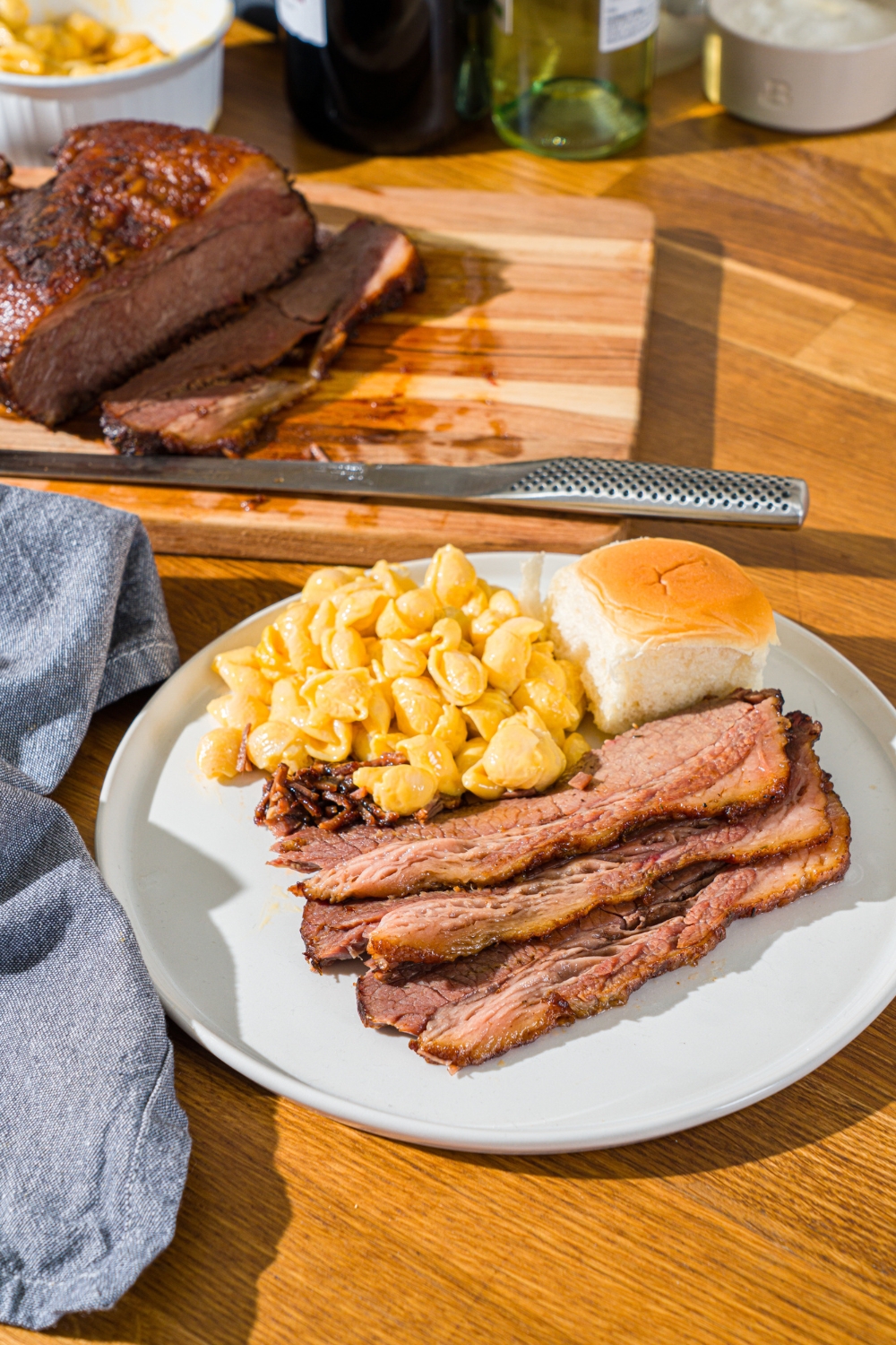 A white plate with sliced oven baked beef brisket, mac and cheese, and a dinner roll. The plate is on a wooden counter with a cutting board of brisket and a blue cloth napkin.