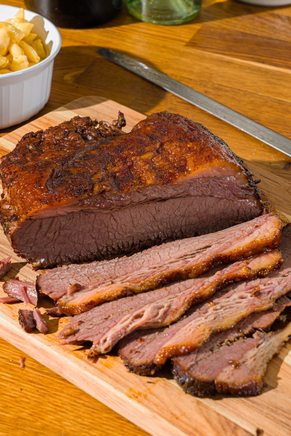 A wooden cutting board with oven baked beef brisket. Some of the brisket has been sliced. There is a bowl of mac and cheese on the board.