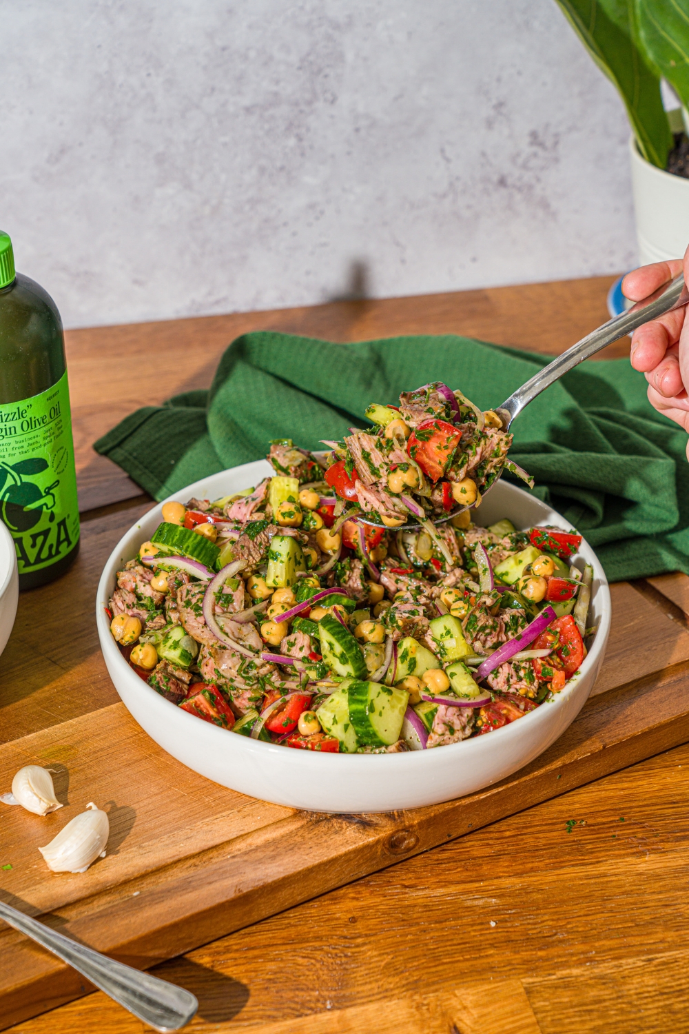 A white bowl with chimichurri steak salad tossed with beans, cucumbers, tomatoes, and red onion in a chimichurri dressing. There is a fork taking a bite from the bowl. The bowl is on a wooden counter with a green cloth napkin.