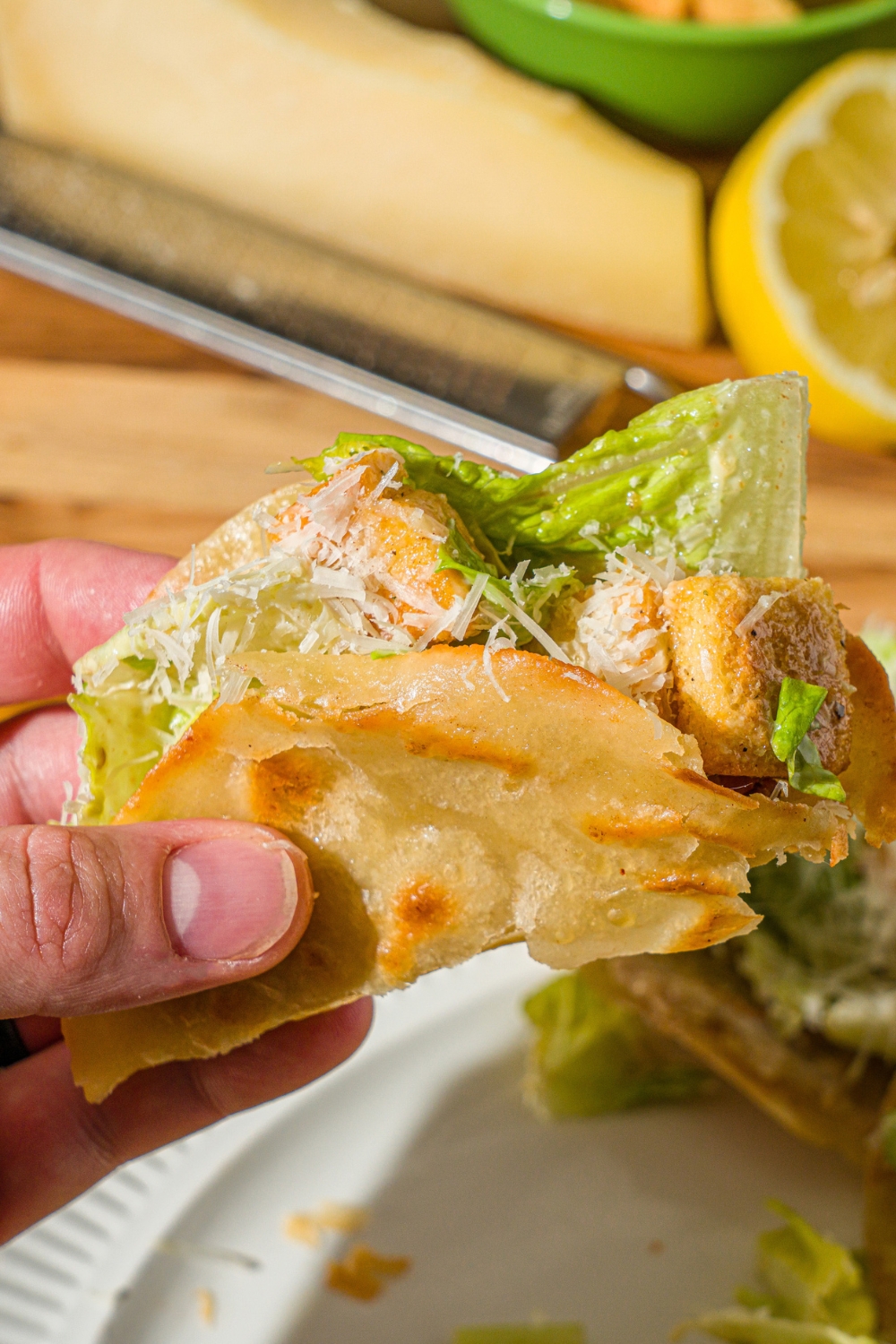 A hand holding a chicken caesar smash taco over a white plate of tacos. The taco is finished with shredded parmesan cheese.