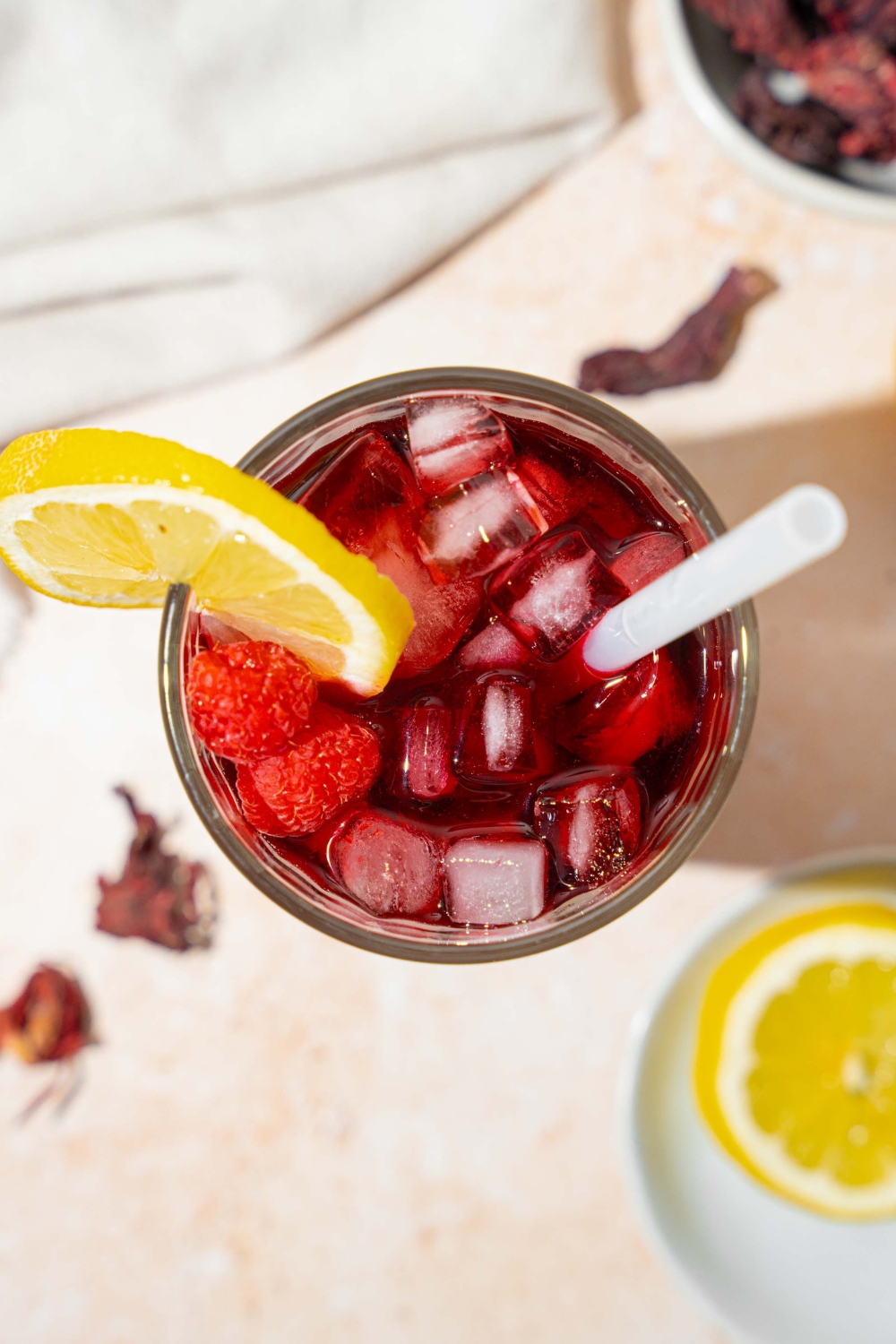 A tall glass of hibiscus lemonade served with ice, sliced lemon, and a straw. The glass is on a tan counter with a white cloth napkin and sliced lemon.