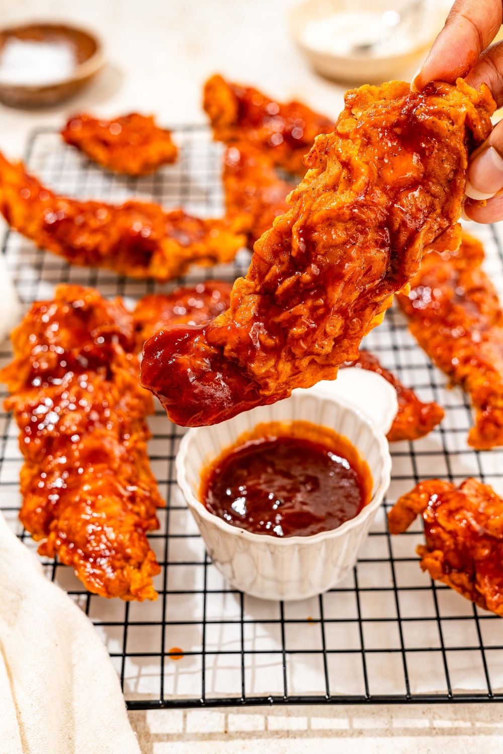 A wire rack with honey bbq chicken tenders and a bowl of honey bbq sauce. one of the tenders is being dipped into the sauce. The rack is on a tan counter.