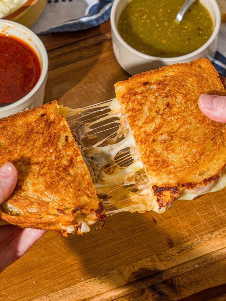 A hand pulling apart a birria grilled cheese sliced in half. There is a wooden cutting board with a bowl of salsa verde and bowl of birria sauce.