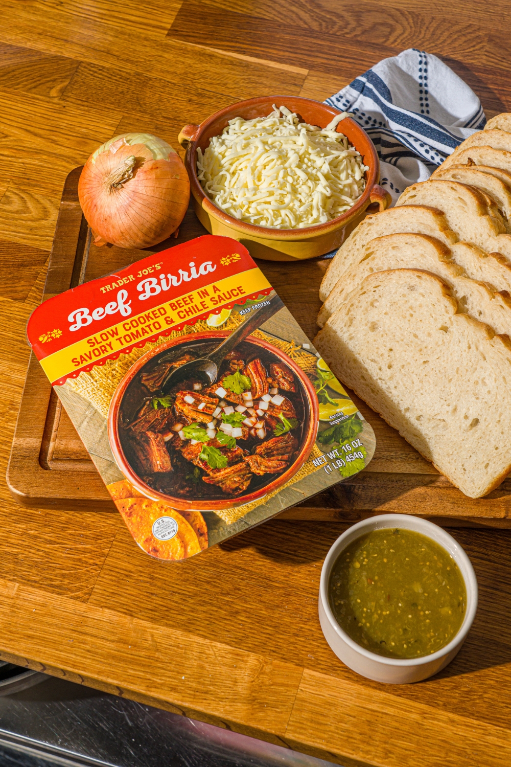 A wooden cutting board with ingredients to make birria grilled cheese including beef birria, shredded mozzarella cheese, salsa verde, onion, and sourdough bread.