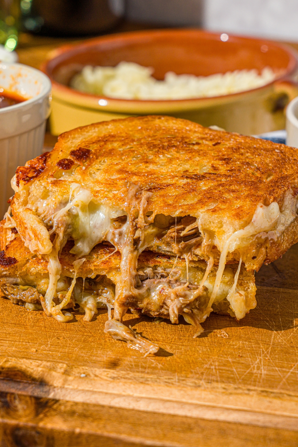 A birria grilled cheese sandwich sliced in half and stacked on one another. The sandwich is on a wooden cutting board with a bowl of birria sauce.