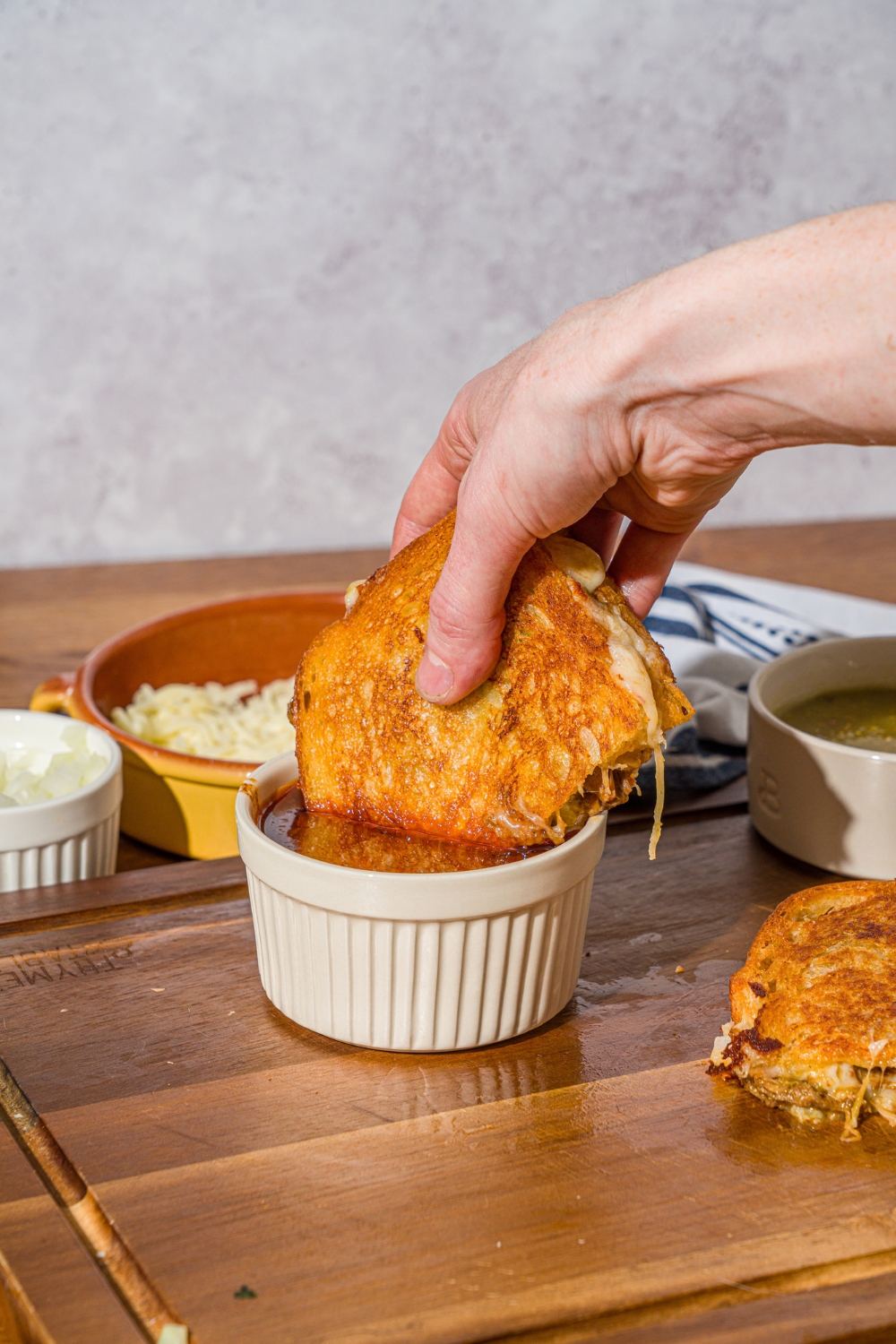 A hand dipping a sliced birria grilled cheese sandwich into a bowl of birria sauce. The bowl is on a wooden cutting board with the other half of the grilled cheese.