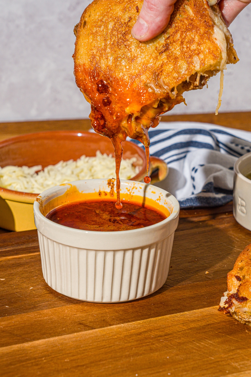 A hand dipping a sliced birria grilled cheese sandwich into a bowl of birria sauce. The bowl is on a wooden cutting board with the other half of the grilled cheese.