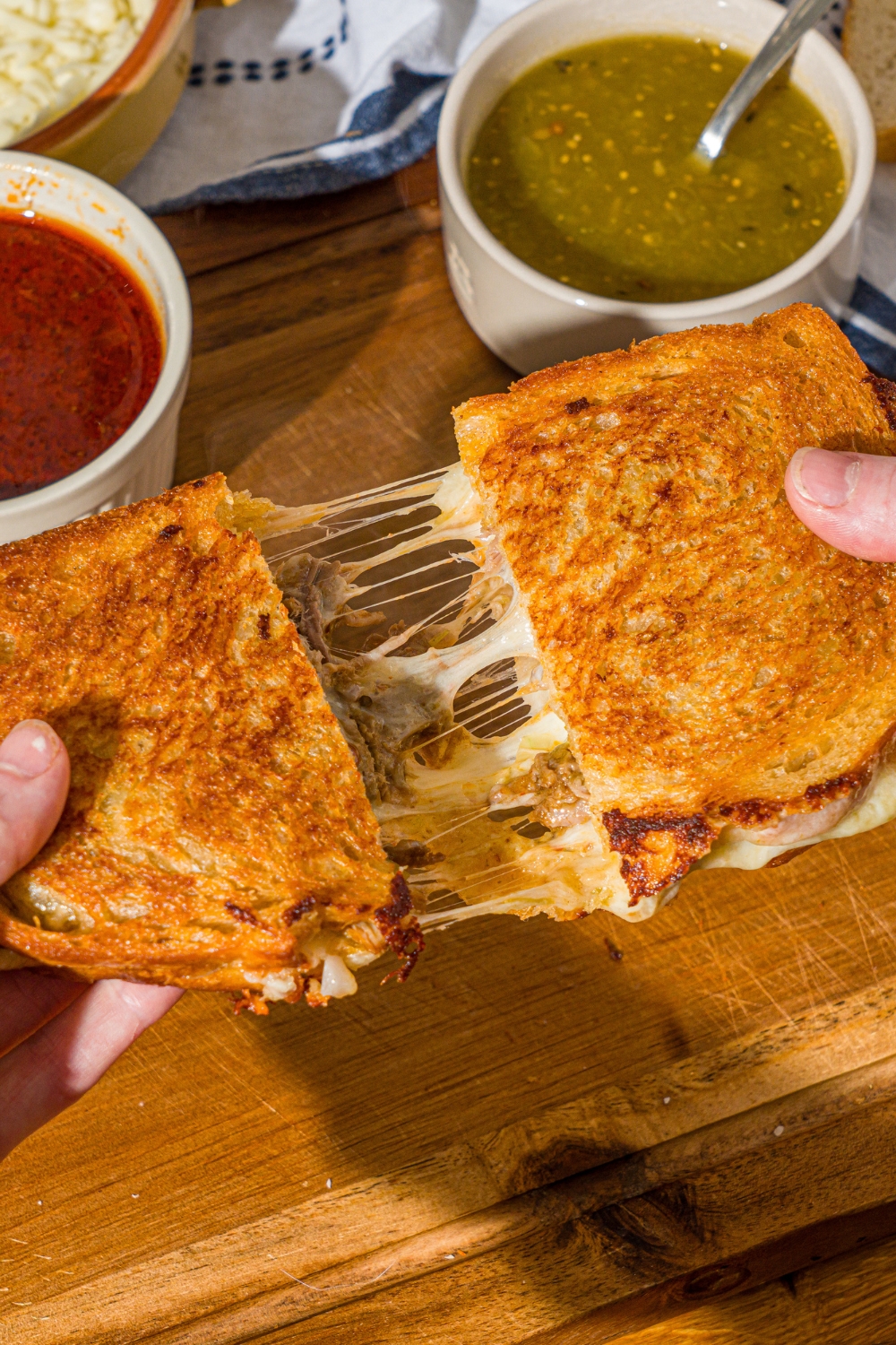 A hand pulling apart a birria grilled cheese sliced in half. There is a wooden cutting board with a bowl of salsa verde and bowl of birria sauce.