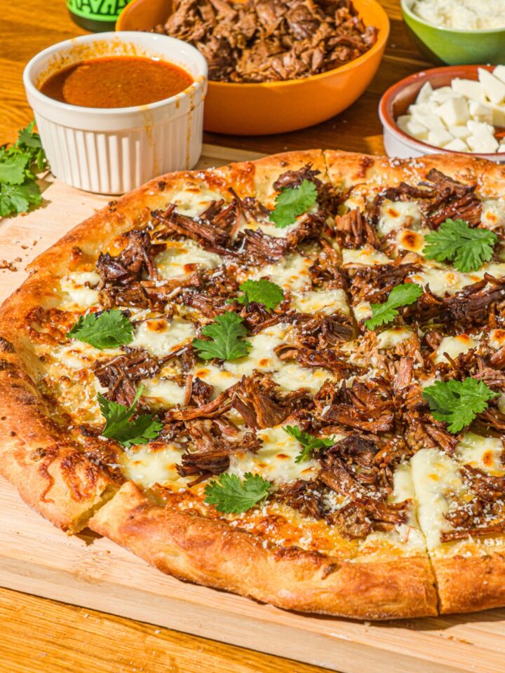 A whole birria pizza topped with fresh cilantro and cotija cheese. The pizza is on a wooden board with a small bowl of mozzarella and birria sauce.