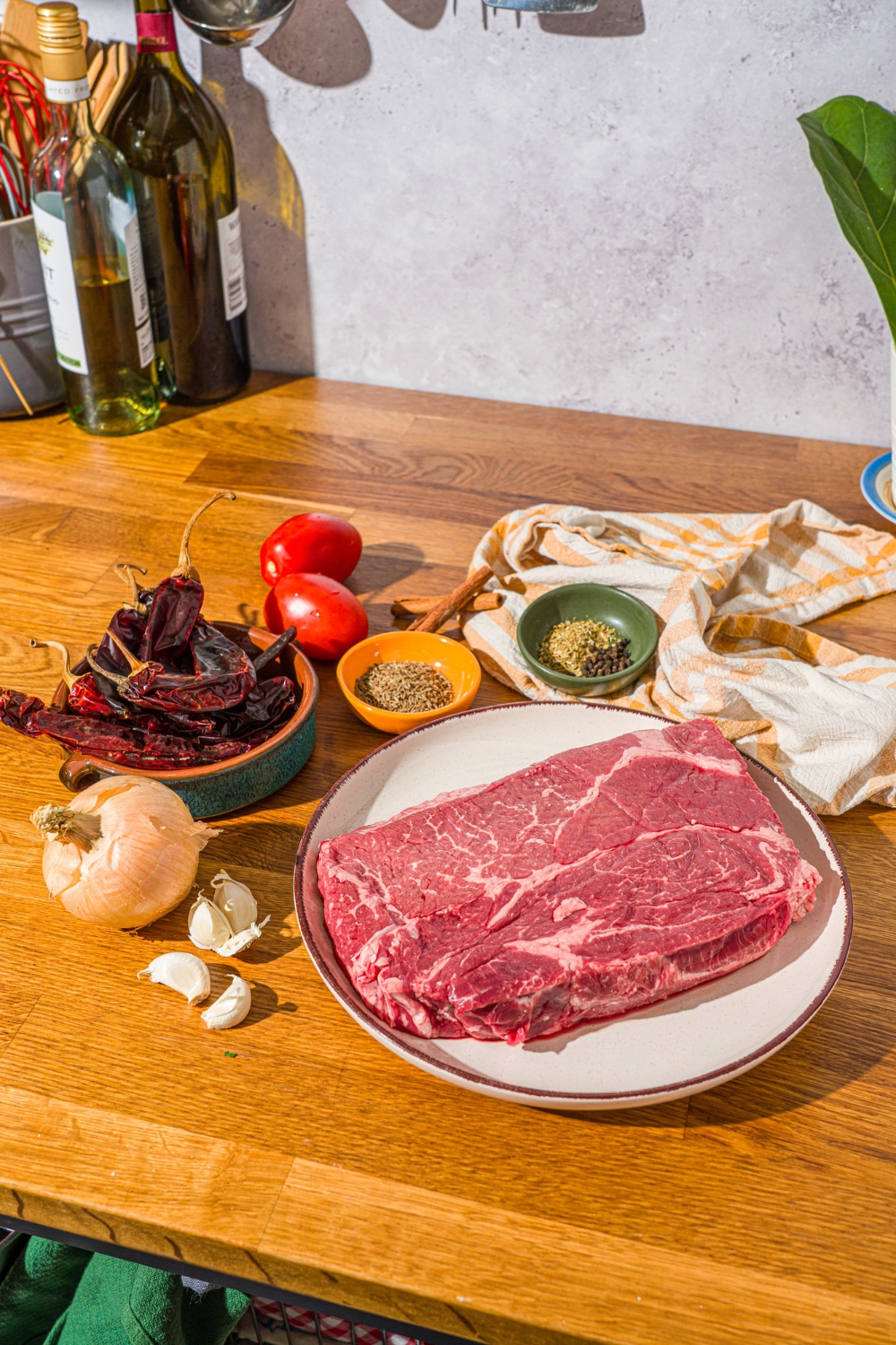 A wooden counter with ingredients to make birria pizza including chuck roast, guajilla peppers, white onion, ancho pepper, roma tomatoes, garlic, and more.