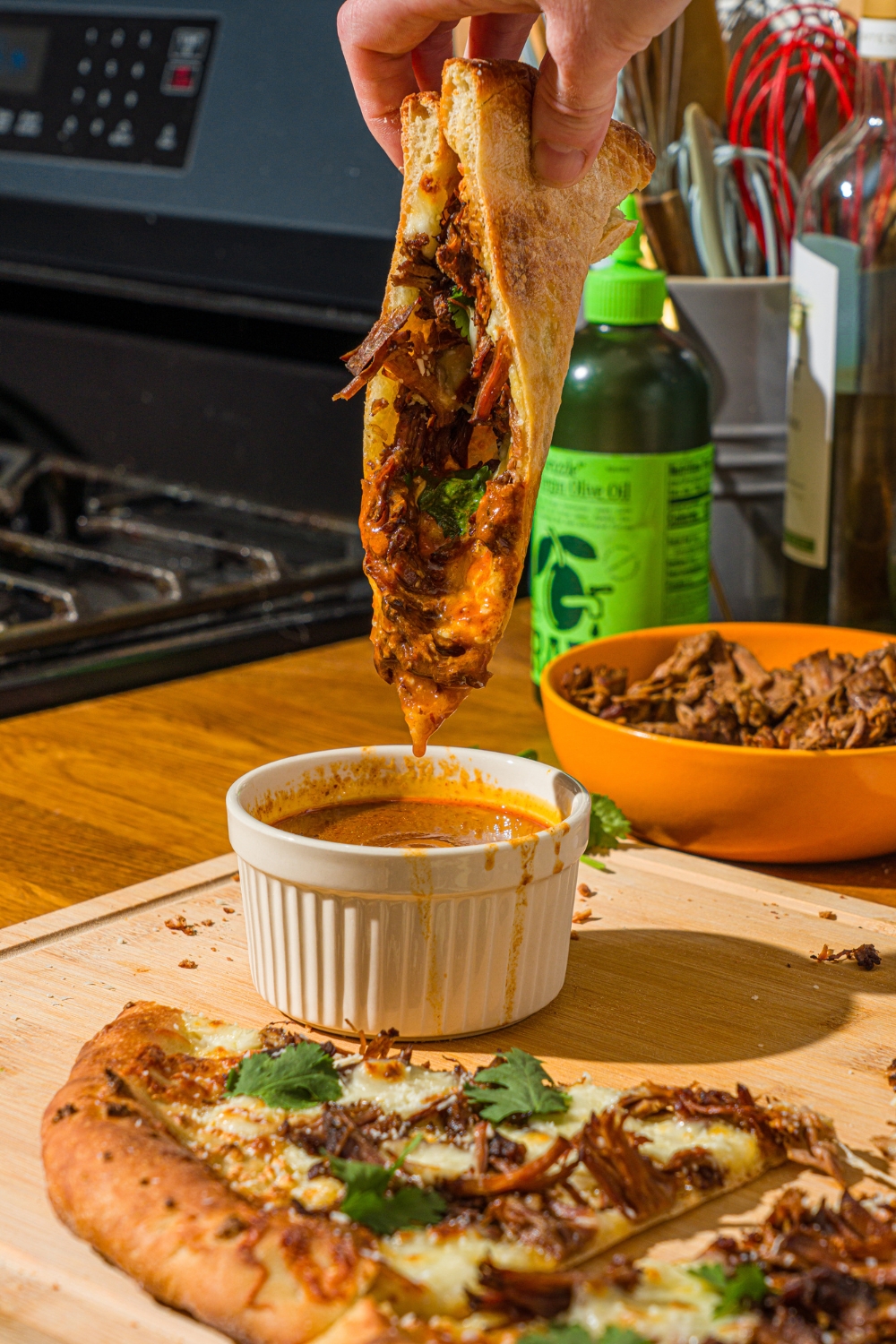 A hand dipping a slice of birria pizza into a bowl of birria sauce. The bowl is on a wooden board with the remaining pizza.