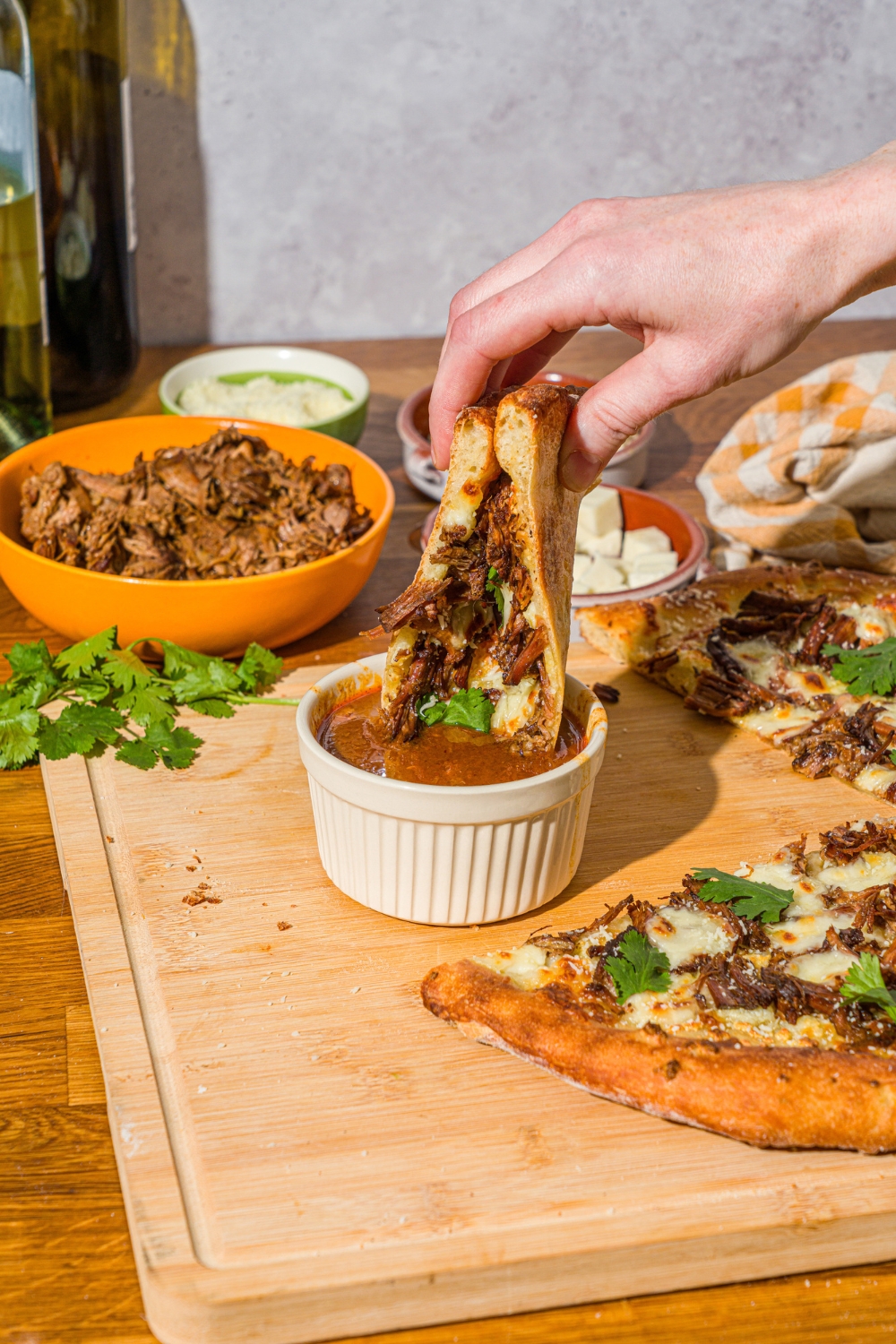 A hand dipping a slice of birria pizza into a bowl of birria sauce. The bowl is on a wooden board with the remaining pizza. The board is on a counter with a bowl of birria meat and mozzarella.