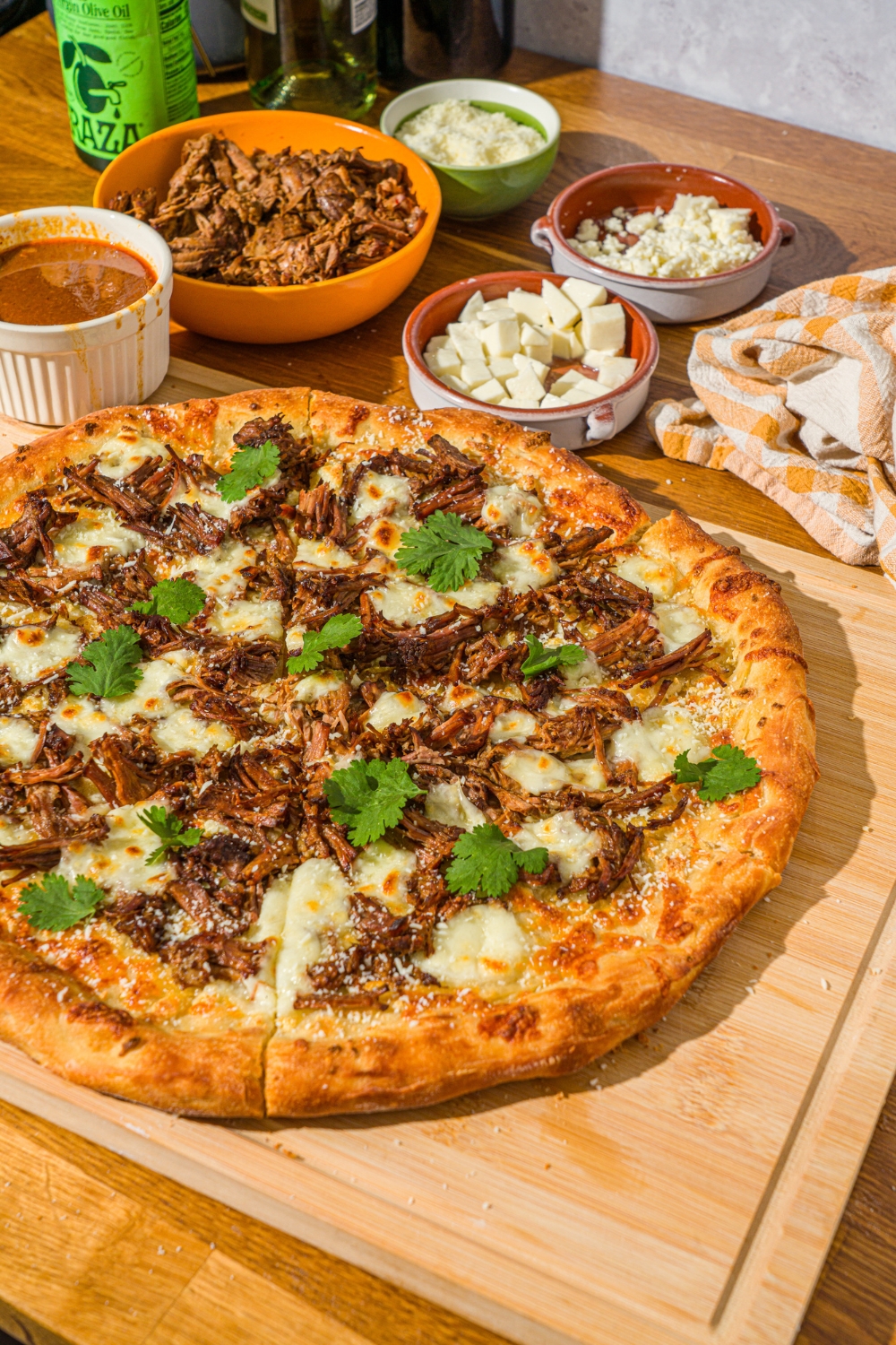 A whole birria pizza topped with fresh cilantro and cotija cheese. The pizza is on a wooden board with a small bowl of mozzarella, cotija cheese, and birria sauce.
