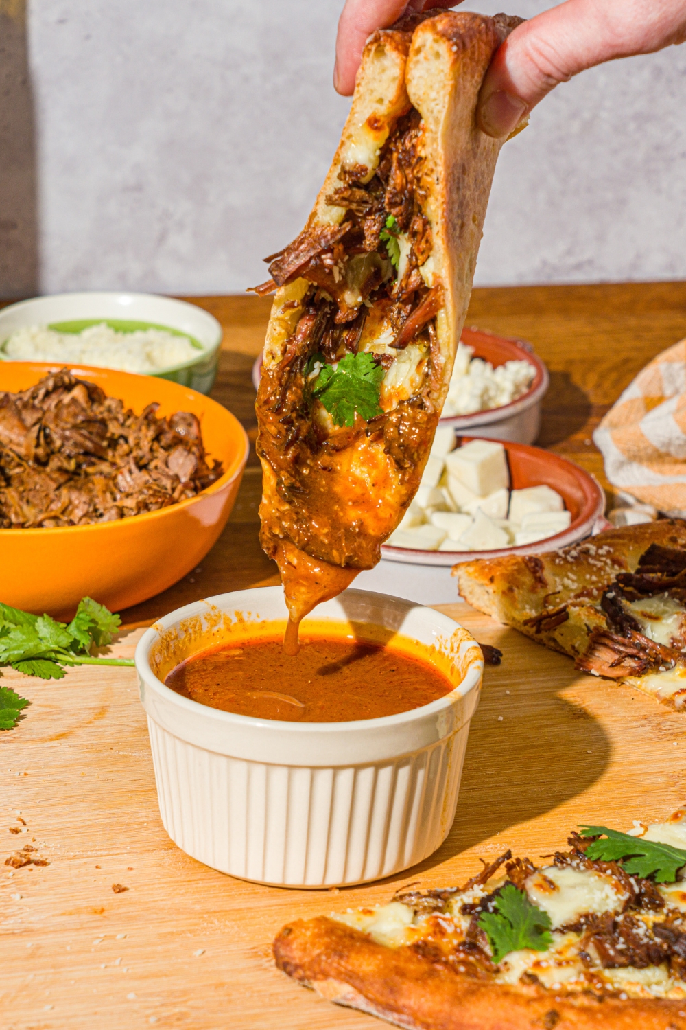 A hand dipping a slice of birria pizza into a bowl of birria sauce. The bowl is on a wooden board with the remaining pizza.