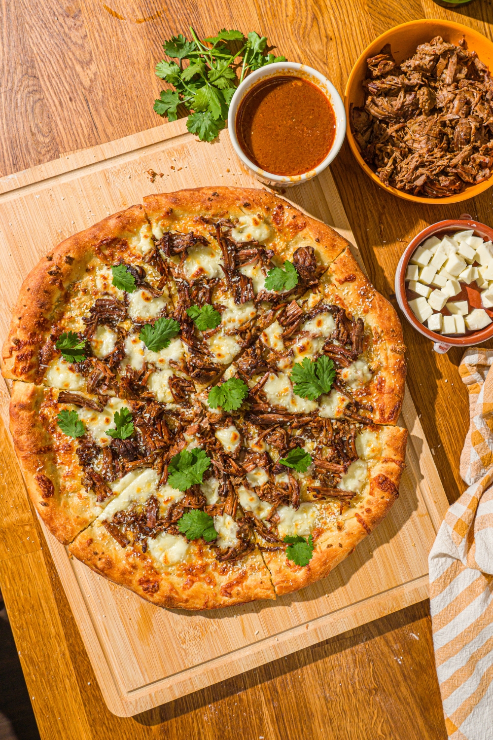 A whole birria pizza topped with fresh cilantro and cotija cheese. The pizza is on a wooden board with a small bowl of mozzarella and birria sauce.