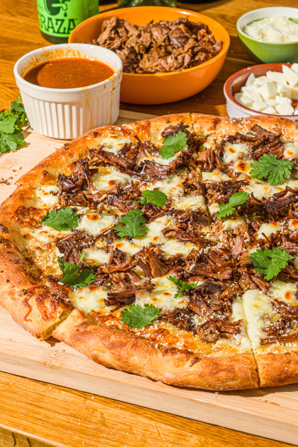 A whole birria pizza topped with fresh cilantro and cotija cheese. The pizza is on a wooden board with a small bowl of mozzarella and birria sauce.
