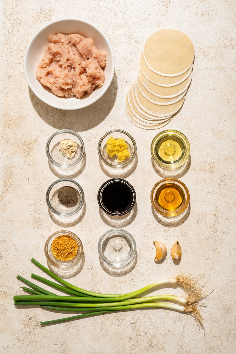 A tan counter with several bowls containing ingredients to make blanket dumplings including dumpling wrappers, ground chicken, garlic, ginger paste, soy sauce, sesame oil, rice vinegar, and green onion.